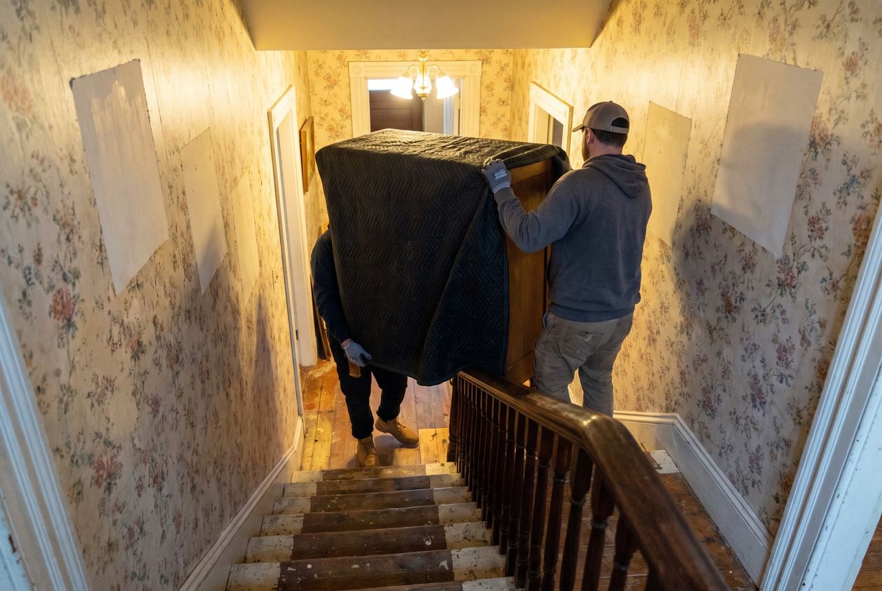 Two movers carrying a blanket-wrapped dresser down a narrow staircase in an older house