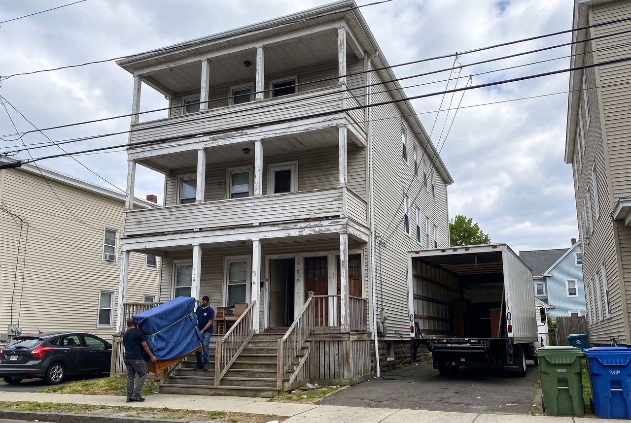 Two movers carrying a blanket-wrapped dresser down the porch stairs of a three-story triple-decker